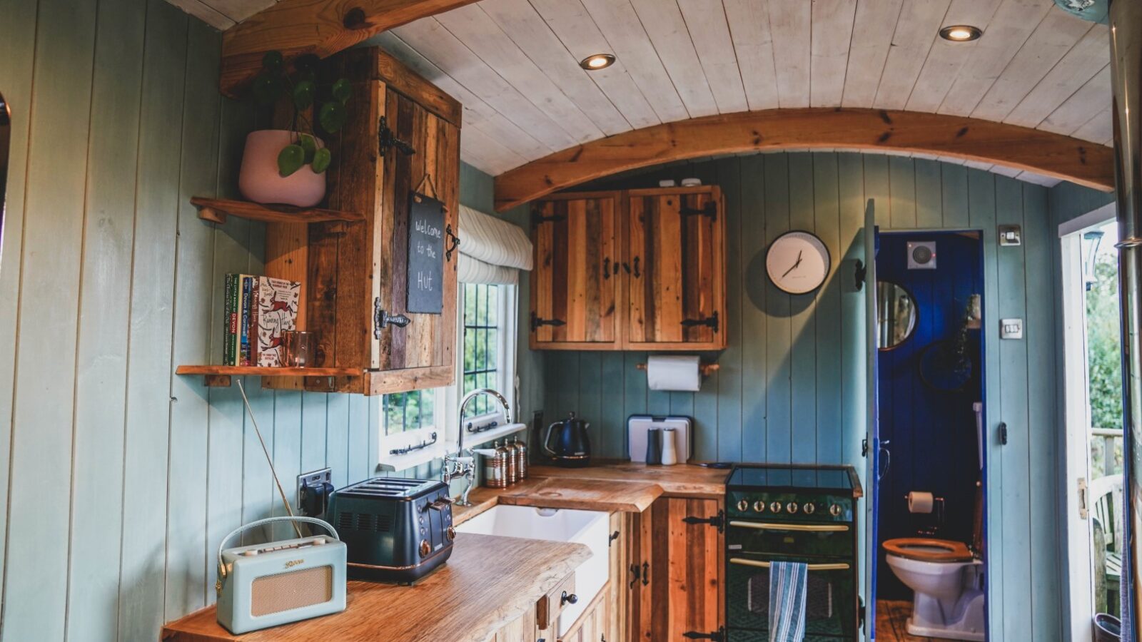 Compact rustic kitchen with wooden cabinets, a gas stove, radio, kettle, and toaster, offering a perfect retreat for those seeking cozy hideaways. The bathroom entrance is visible on the right. Could easily be part of the charming ambiance at Leworthy Manor.