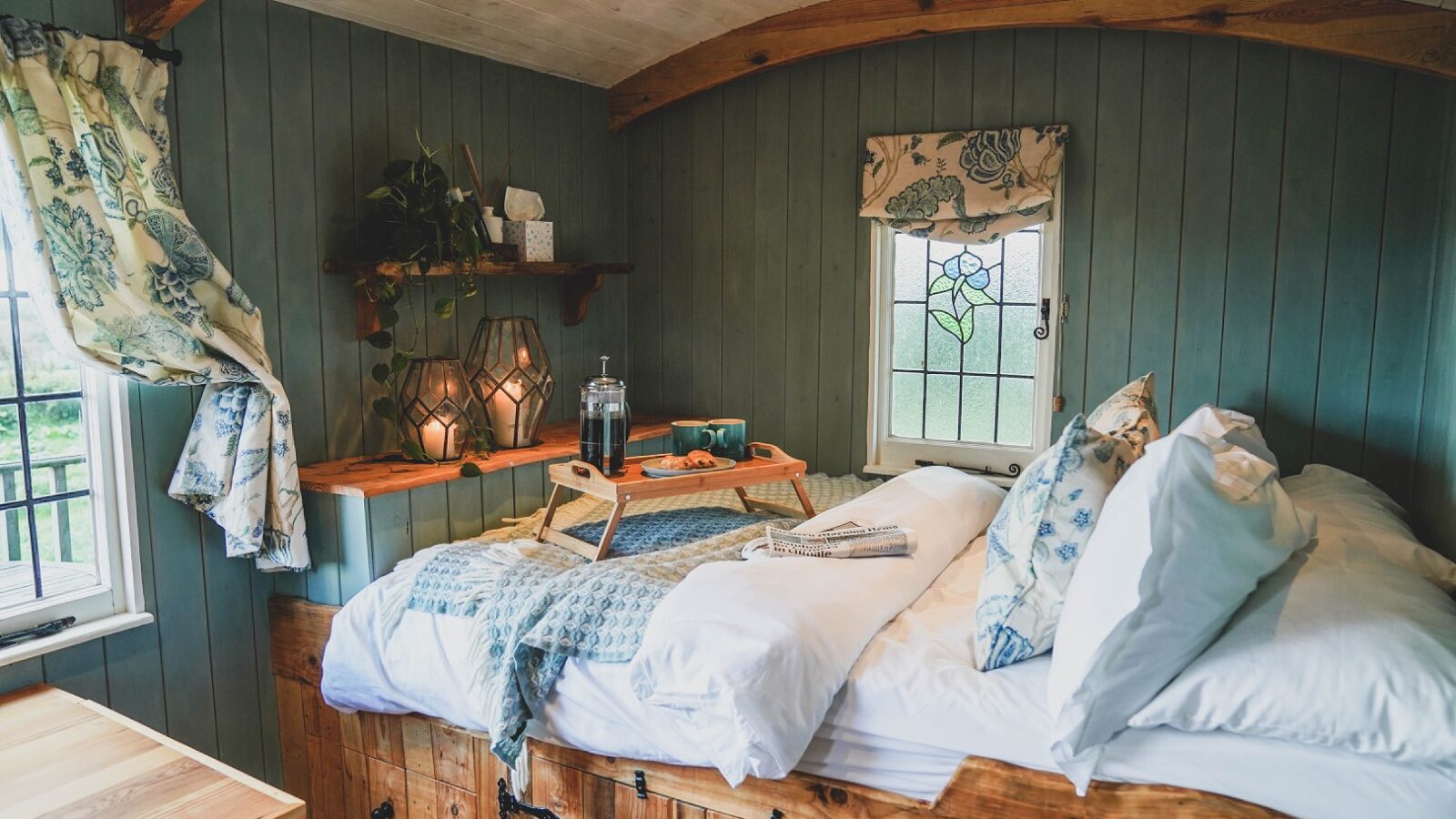 Cozy bedroom at Leworthy Manor features a wooden bed, blue-green walls, decorative pillows, a tray with drinks, and a window with stained glass—ideal for serene hideaways.