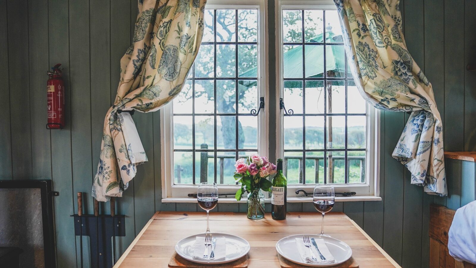 A cozy dining setup for two at Leworthy Manor, featuring wine and flowers on a wooden table by a window with floral curtains and an enchanting outside view, perfect for intimate hideaways.