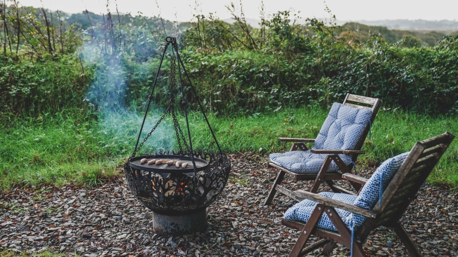 Two cushioned wooden chairs face a decorative fire pit with smoke, set on a stone patio surrounded by greenery, offering a serene escape at Leworthy Manor. Perfect for those seeking tranquil hideaways amidst nature's embrace.