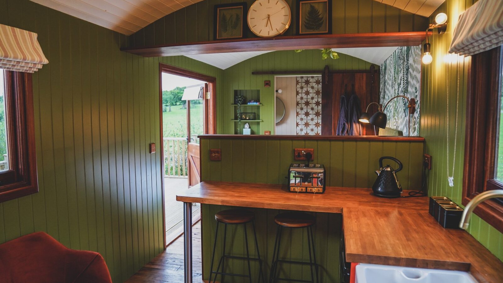 Cozy cabin kitchen with green walls, wooden counter, black kettle, bar stools, and wall clock, offering a serene view reminiscent of the lush fields surrounding Leworthy Manor hideaways.