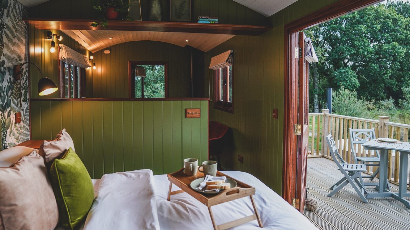 Cozy cabin interior at Leworthy Manor, featuring a bed and breakfast tray. An open door leads to a deck with chairs, offering serene hideaways surrounded by trees.