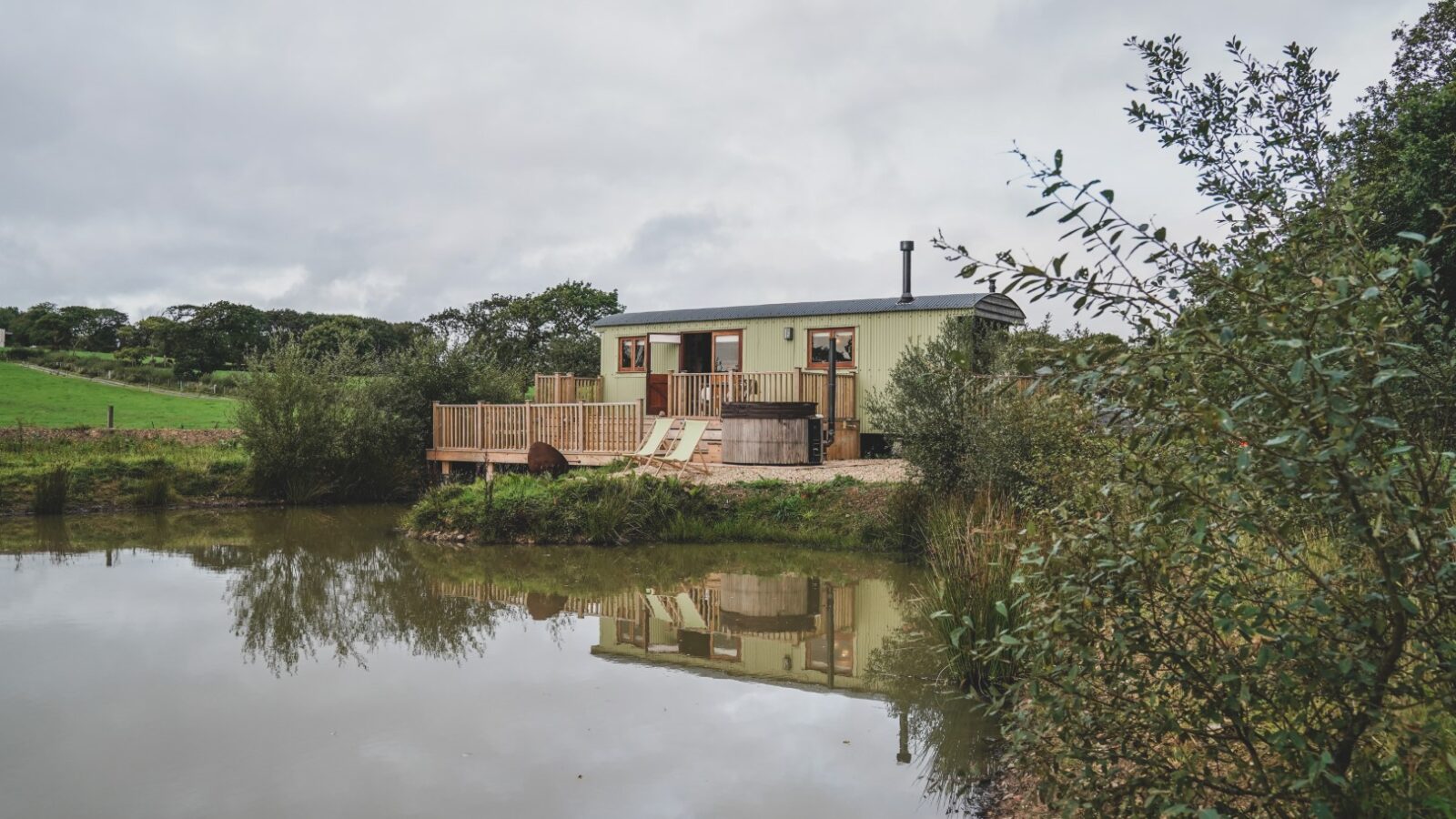 Nestled amidst trees and grass, a small cabin from Leworthy Manor's Hideaways offers a deck that overlooks a tranquil pond under a cloudy sky.