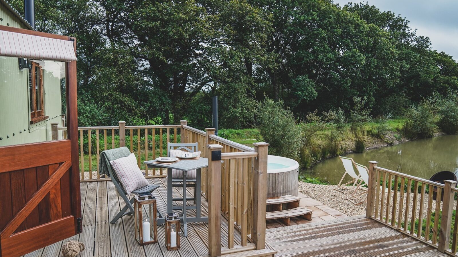 The wooden deck at Leworthy Manor features a cozy chair, table, and lanterns, offering a serene escape. Overlooking a hot tub and lush greenery near the pond, it's one of the most charming hideaways for relaxation.