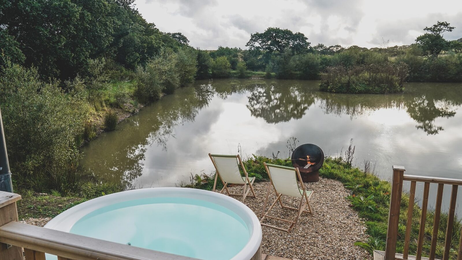 Nestled in the serene landscape of Leworthy Manor, this outdoor patio features two chairs by a tranquil pond, a small pool, and a cozy fire pit. It's an idyllic hideaway surrounded by lush greenery under a cloudy sky.