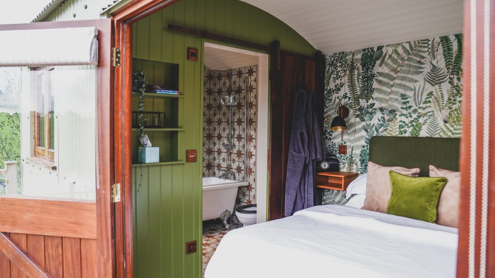 Cozy bedroom with a green theme, reminiscent of the charming Leworthy Manor, opens to reveal a small bathroom adorned with patterned wallpaper and a clawfoot bathtub—a true hideaway.