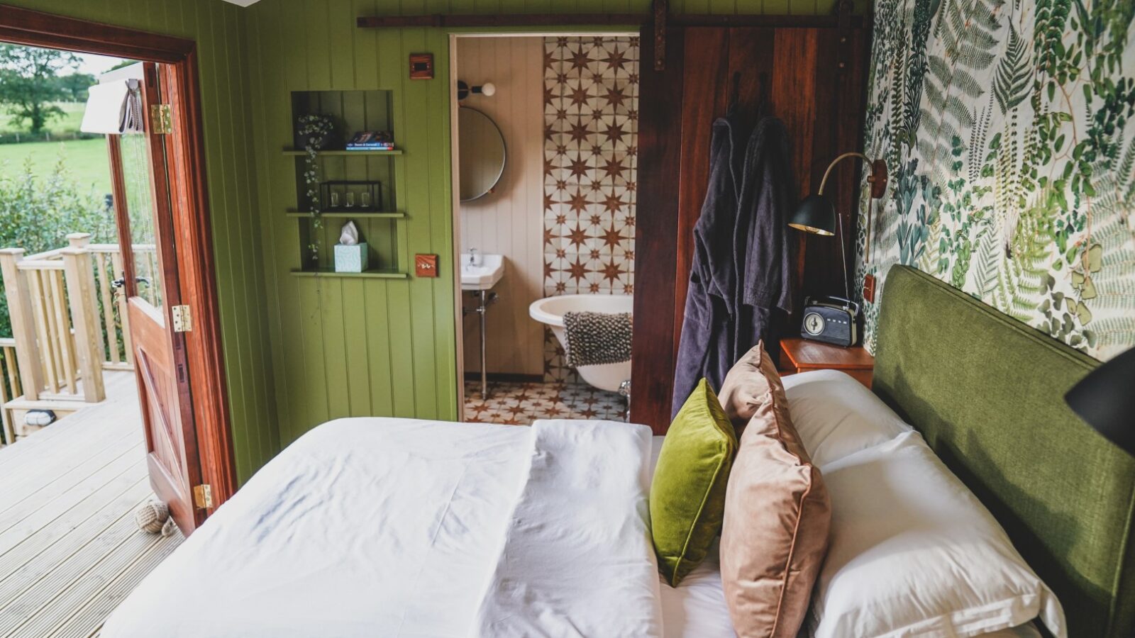 Cozy bedroom at Leworthy Manor Hideaways featuring a double bed, green decor, open door to a deck, and view of a bathroom with patterned tiles.