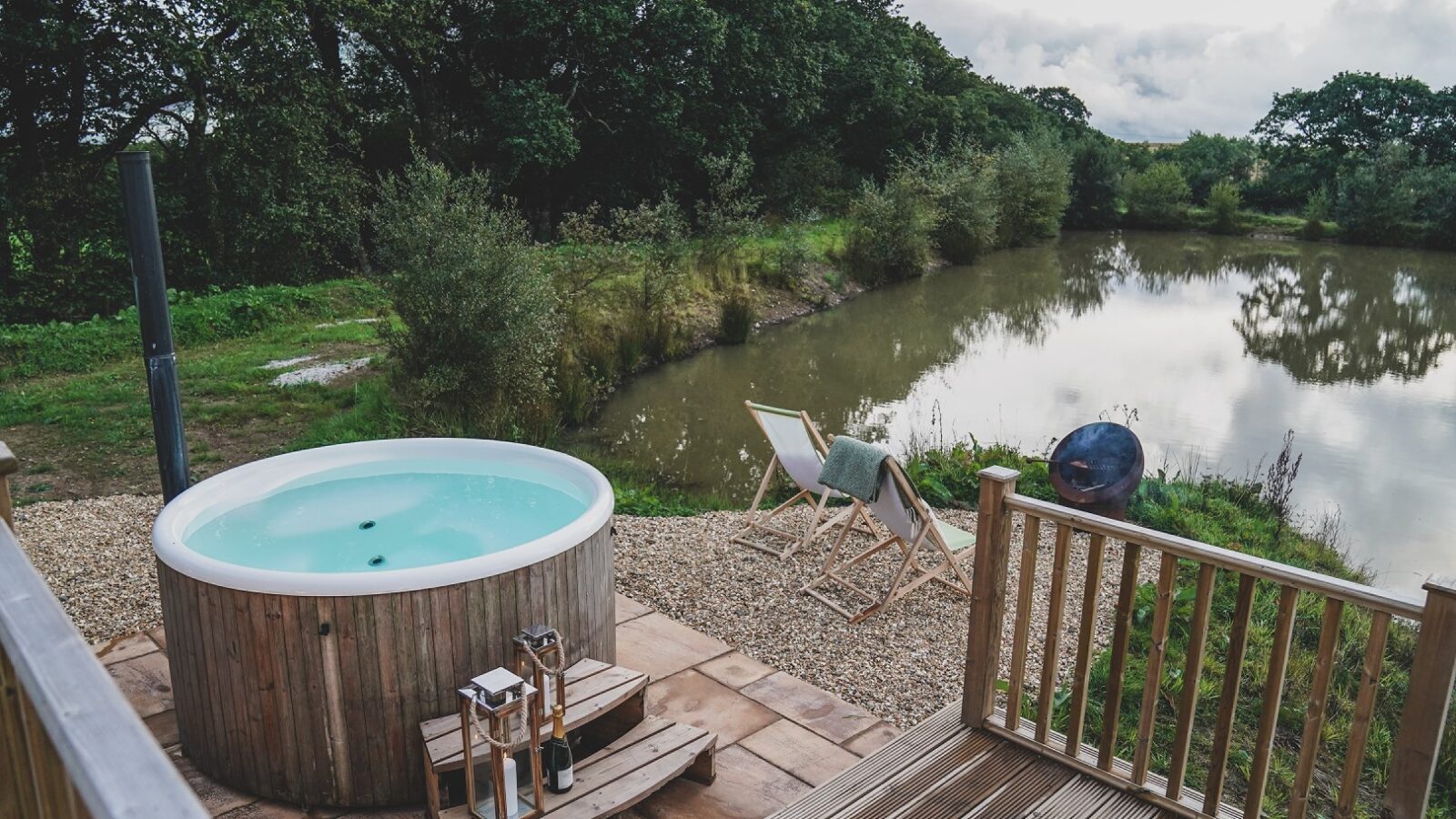 Nestled within the scenic Hideaways of Leworthy Manor, this outdoor deck features a round hot tub, two cozy chairs, and a small table overlooking a tranquil pond surrounded by lush greenery.