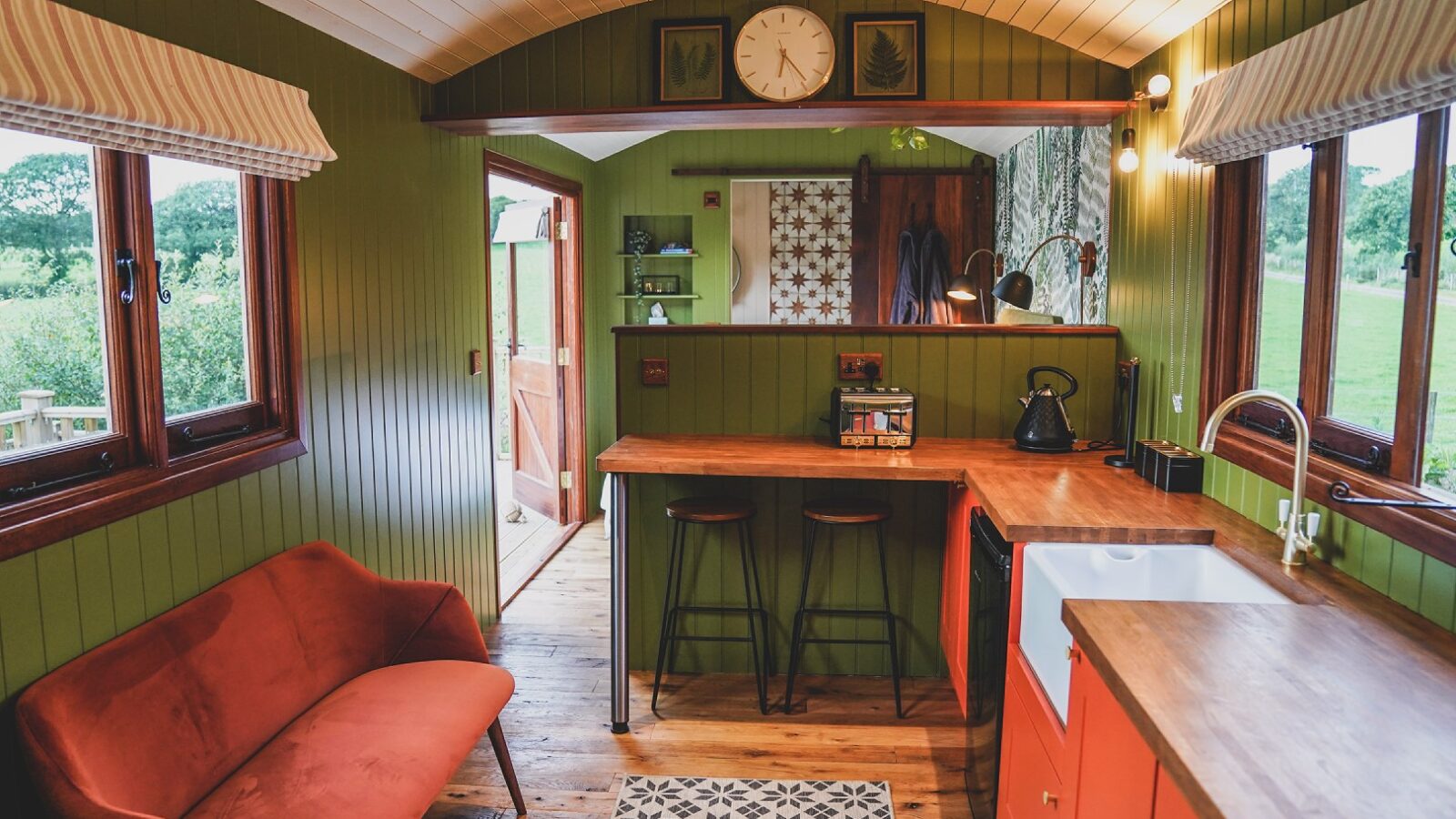 The cozy cabin interior at Leworthy Manor Hideaways features green walls, a wooden countertop, and vibrant orange accents. A red couch complements the view of lush greenery outside, creating a perfect retreat.