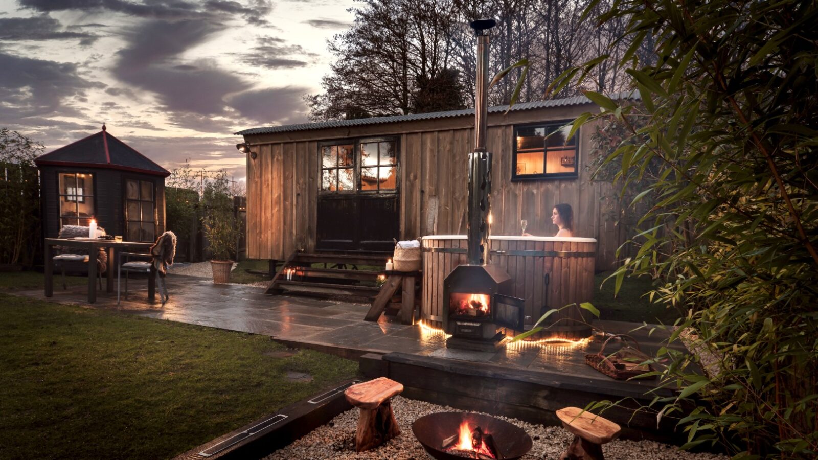 A cozy wooden retreat at dusk features a hot tub and lit fire pit, with a small dining area nearby offering chairs and a table for relaxing evenings under the stars.