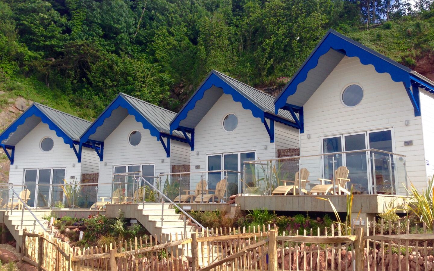 Four identical beach huts with blue roofs and white walls, situated on a hillside amid lush greenery, and enclosed by a wooden fence in front.