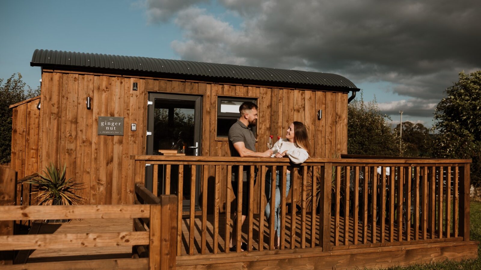 Two people stand on a wooden deck in front of a cozy shepherd's hut, surrounded by greenery and under a partly cloudy sky.