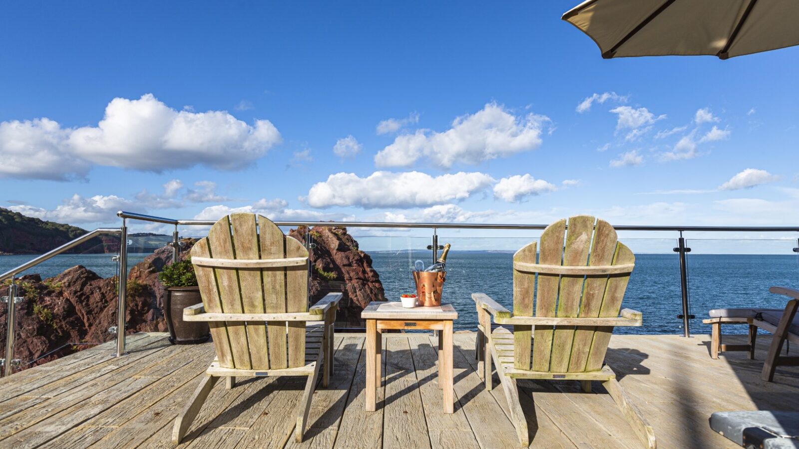 Two wooden chairs on the wooden deck of a Cary Arms suite overlook the ocean under a clear blue sky, with a table and plant nestled between them.