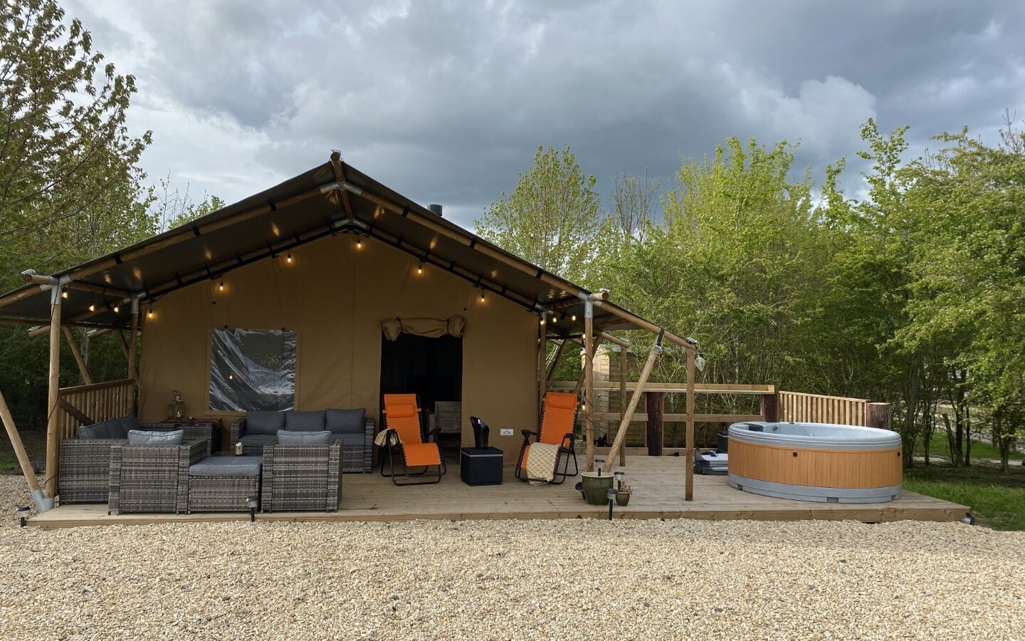 A glamping safari tent with orange chairs and a seating area on a wooden deck, located next to a hot tub. The cloudy sky overhead adds to the charm of Chilterns View.