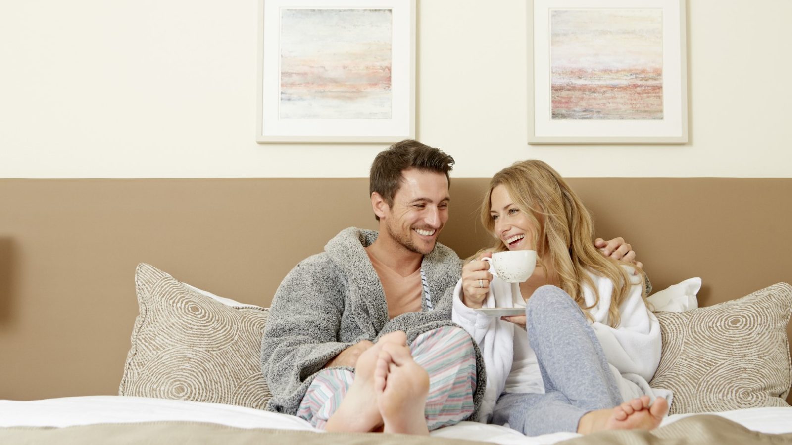 A couple sitting on a bed in cozy attire, laughing and holding a cup of coffee. The room evokes the warmth of a barn with soft lighting, neutral colors, and two abstract paintings above the headboard. Both appear relaxed and happy in this intimate retreat.