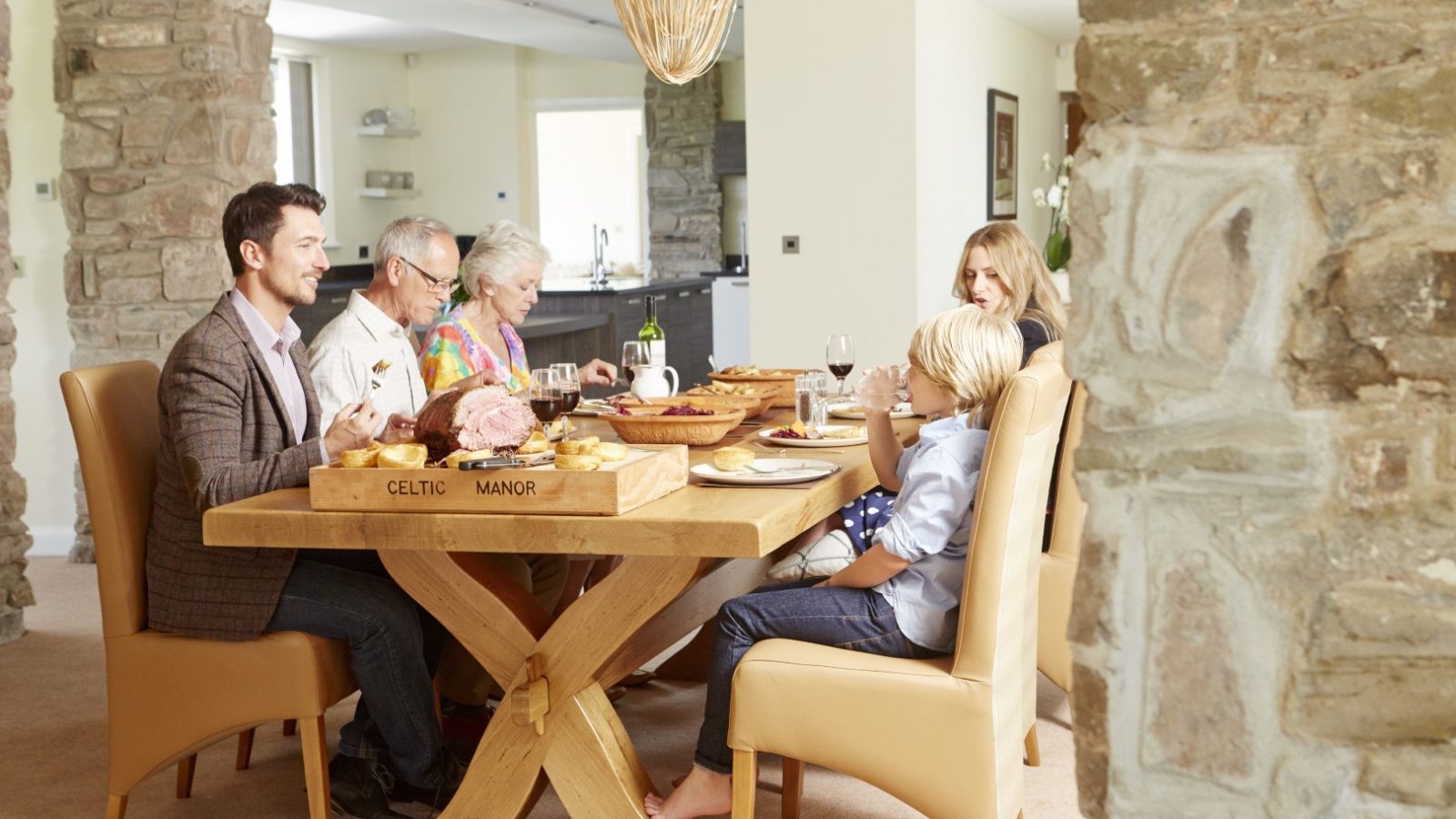 A family of six gathers around a wooden dining table for a meal in their cozy home. Reminiscent of a Celtic Manor, the table is set with dishes, and everyone enjoys each other's company against stone and cream-colored walls.