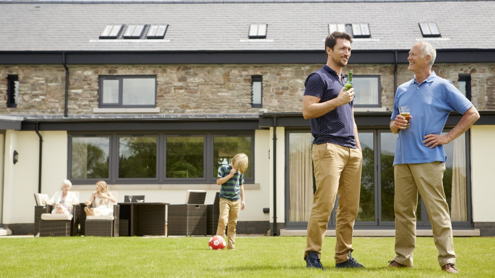 In the garden of a modern house reminiscent of the Celtic Manor style, two men stand sipping drinks and chatting. A child kicks a soccer ball nearby, while two others relax on a patio set. The scene is bright and cheerful.