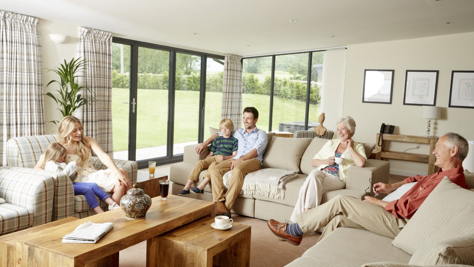 A family gathers in a bright living room with large windows, reminiscent of the cozy charm of a Celtic Manor Barn. Two adults sit with a child on the couch, another adult holds a child in an armchair, and two older adults relax on the opposite couch. The space is decorated with plants and framed art.