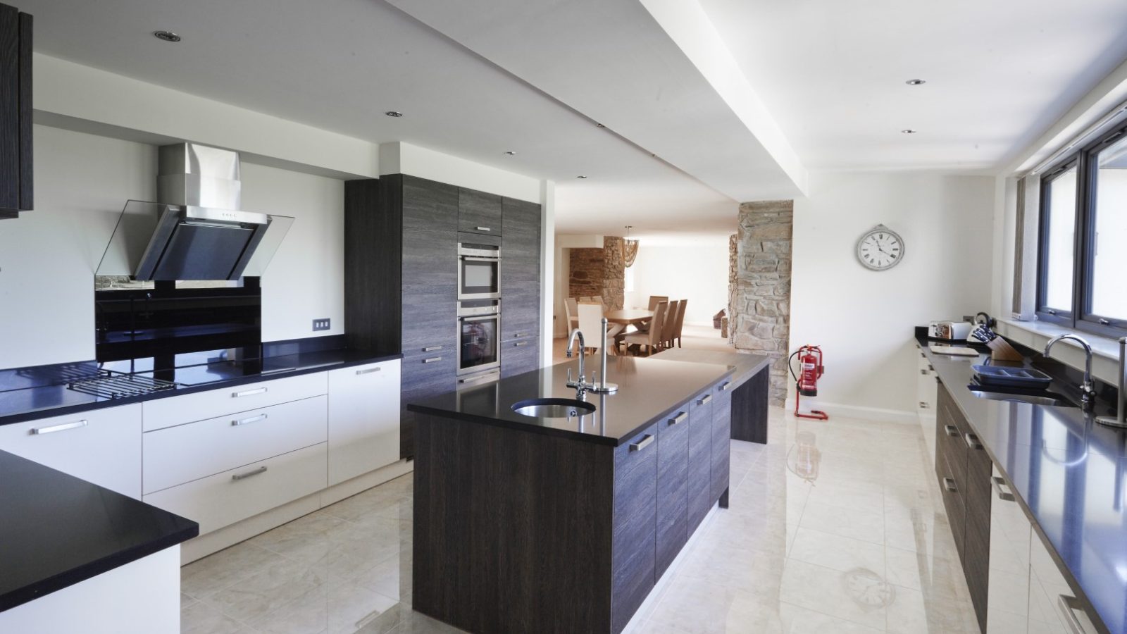 Modern kitchen with sleek design, featuring dark wood cabinets, white drawers, and black countertops reminiscent of a refined Celtic Manor. A central island with a stove and chimney hood. Large windows provide natural light to the dining area, where a wall clock is visible in the background.