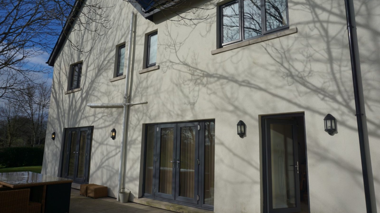 A two-story farmhouse with cream-colored walls and several windows, casting tree branch shadows. Glass double doors lead to a stone patio with outdoor seating. The sky is clear and blue, reminiscent of a serene day at Celtic Manor.