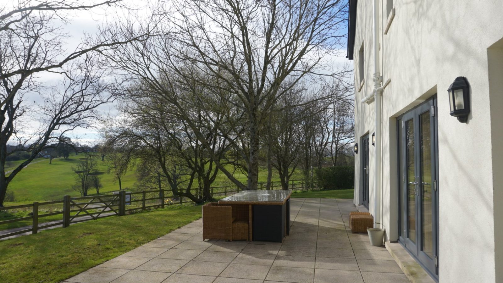 A charming patio with a glass table and wicker chairs sits beside a large white farmhouse. Bare trees and a grassy field stretch out under the partly cloudy sky, adding to the serene atmosphere.