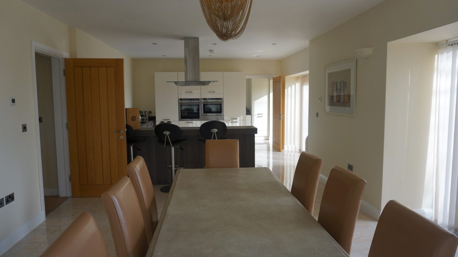 In a blend of modern and farmhouse charm, the kitchen and dining area features a long table surrounded by brown chairs. A Celtic Manor vibe is evoked with the island's high stools, oven, and range hood. A large chandelier crowns the scene while sliding glass doors drench it in natural light.