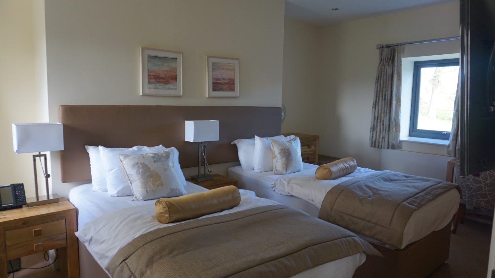 A cozy hotel room at Celtic Manor features two neatly made twin beds with white linens, decorative pillows, and bed runners. Two lamps rest on a shared bedside table. Natural light spills through the window on the right, while farmhouse-style framed art adorns the wall above the beds.