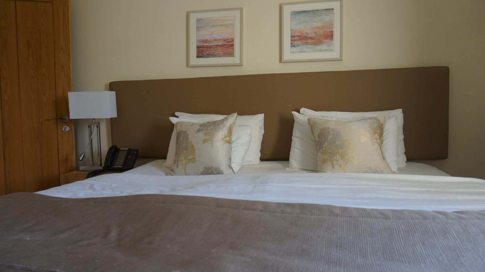 A cozy hotel room with a neatly made double bed, featuring beige and white bedding reminiscent of a farmhouse style. Two decorative pillows with a tree motif add charm. Above the bed, two framed abstract paintings hang on the light-colored wall. A phone and lamp are on the bedside table, adding to its Celtic Manor allure.
