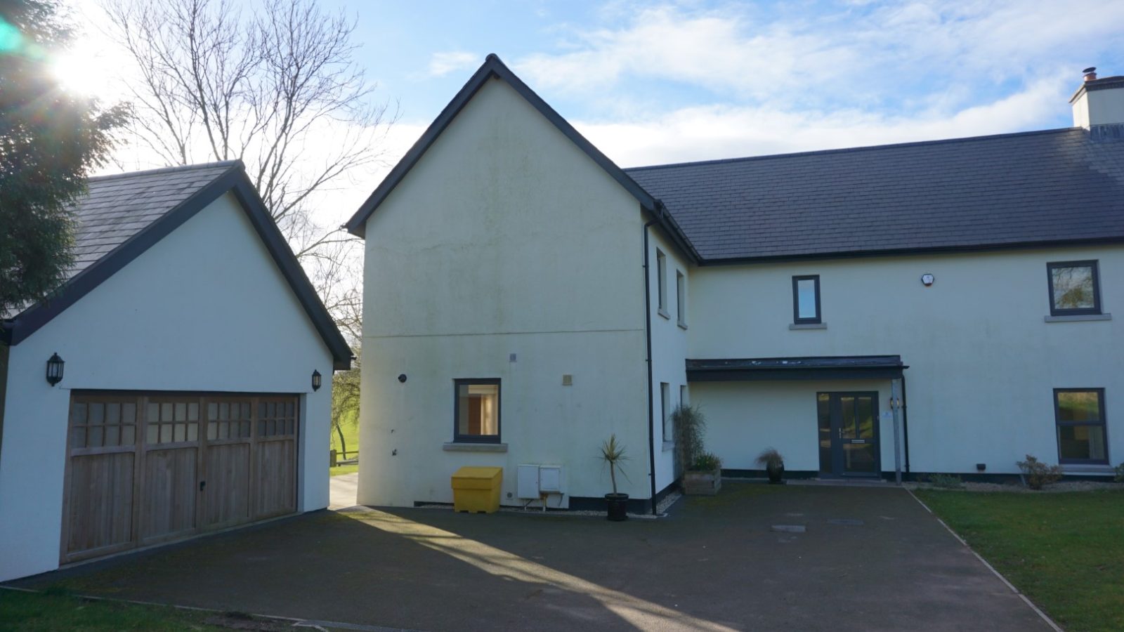 A modern two-story farmhouse with a gray roof, white walls, and several windows stands beside a two-car garage with wooden doors. The property, reminiscent of a Celtic Manor, sits on a spacious driveway surrounded by trees under a cloudy blue sky.