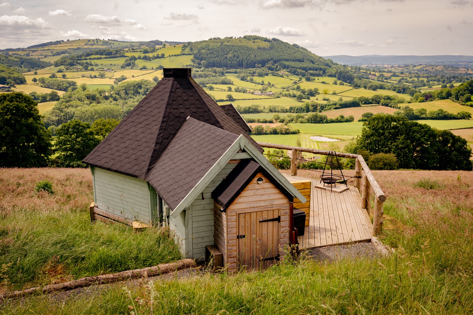 Experience unique stays in the UK with this charming wooden cabin, featuring a shingled roof and nestled in a grassy field. It offers breathtaking views of rolling hills and scattered trees under a partly cloudy sky.