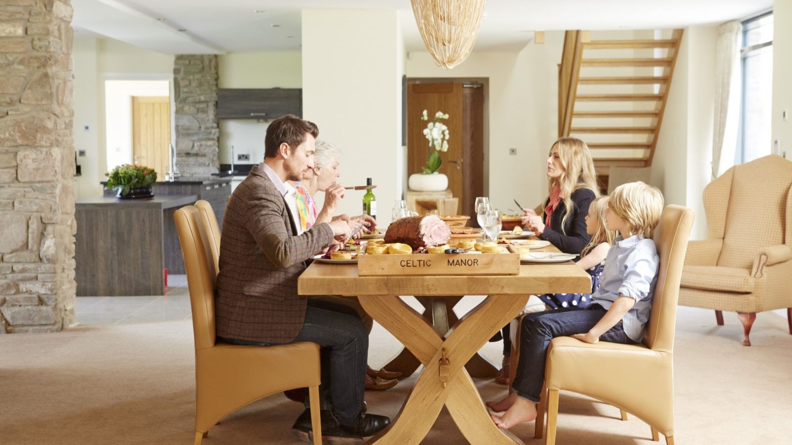 A family of five, including three adults and two children, is seated around a dining table enjoying a meal in the spacious, modern dining room of their Hunter Lodge at Celtic Manor. The table is labeled 