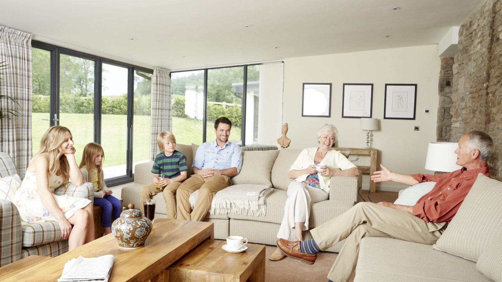 In a modern living room with large windows, a group of six people, including three children and three adults, are sitting and chatting. The cozy space resembles the warmth of Hunter Lodges. A coffee table with cups and decor graces the foreground, while a stone wall rests on the right.