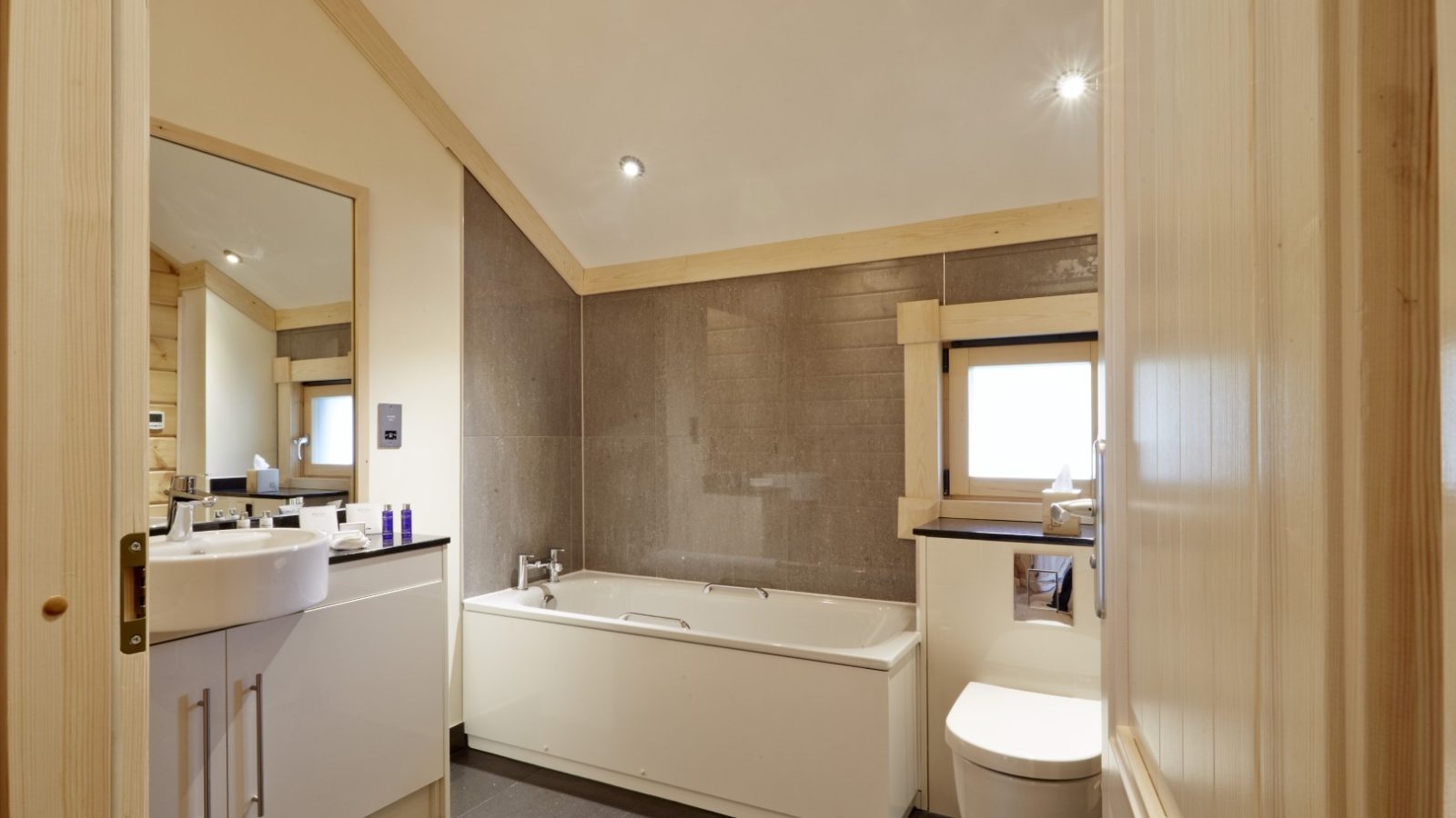 A modern bathroom in the style of Hunter Lodges, with a beige and gray color scheme. It features a bathtub, wall-mounted sink with a large mirror, toilet, and small window. Ceiling spotlights offer ample lighting, while wooden accents on walls and ceiling add rustic charm.