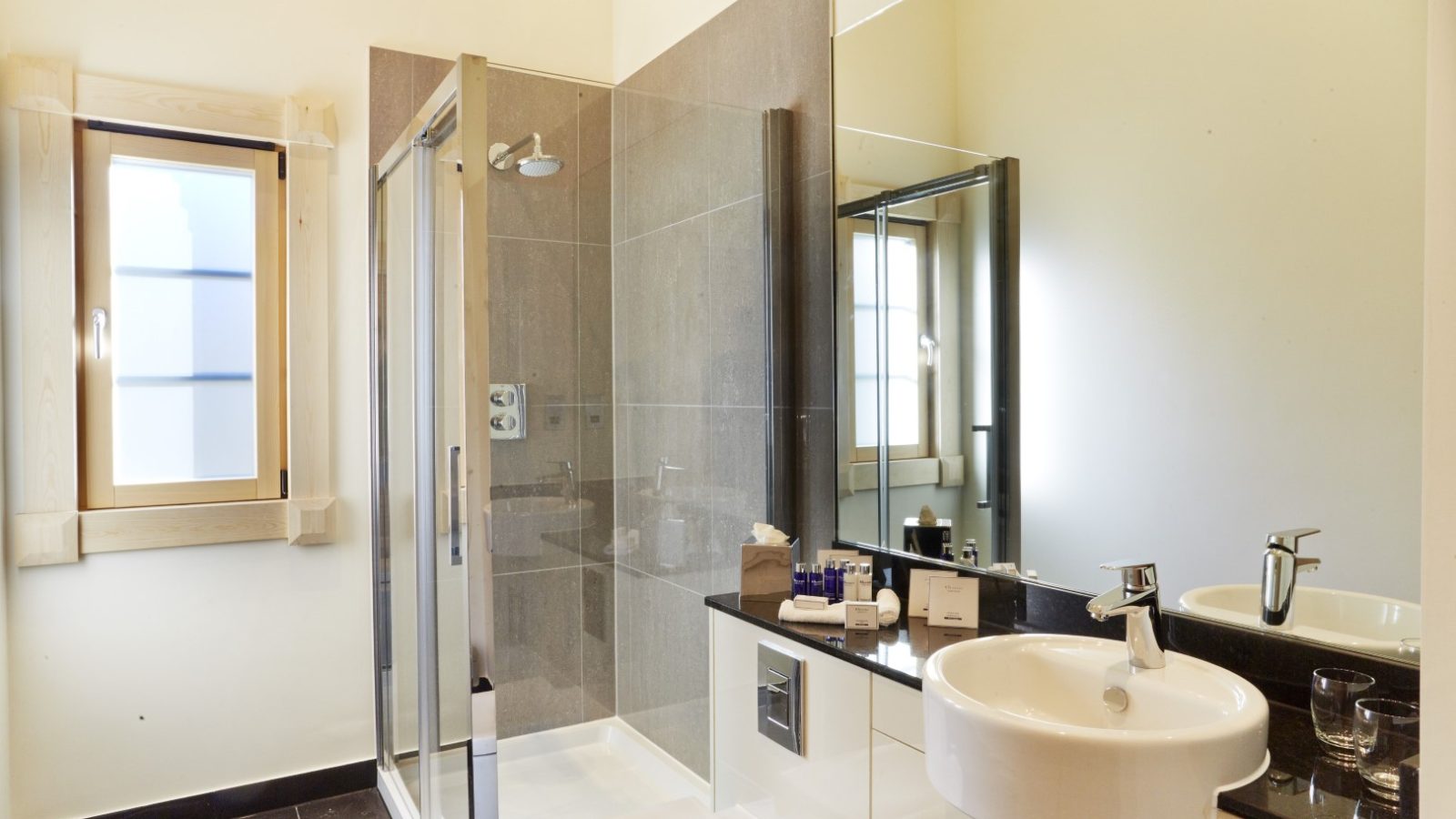 A modern bathroom at Celtic Manor's Hunter Lodges features a glass-enclosed shower, a white sink, and a mirror above the countertop. Natural light streams through the window, illuminating the space where various toiletries are neatly arranged.