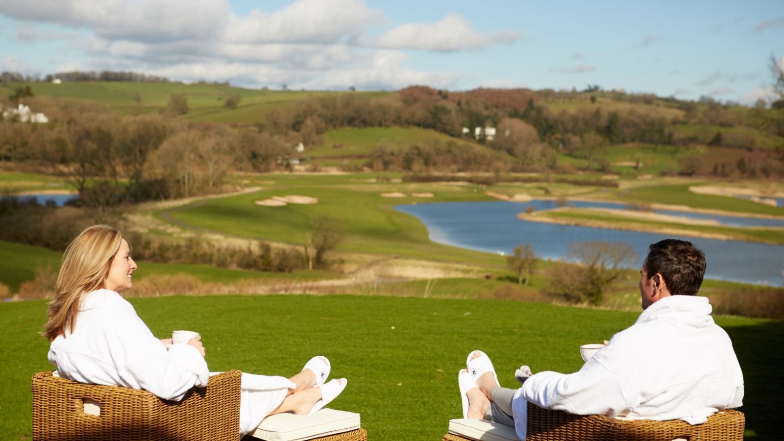 Two people in white robes relax in wicker chairs at Hunter Lodges, facing a scenic view of a lush, green landscape with a winding river and gentle hills under a blue sky. They appear to be enjoying a peaceful moment outdoors.