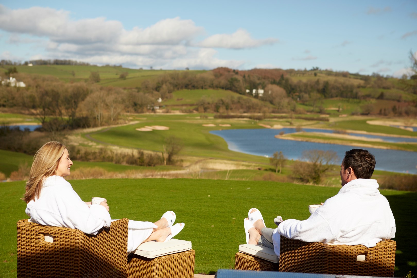 Two people in white robes relax in wicker chairs at Hunter Lodges, facing a scenic view of a lush, green landscape with a winding river and gentle hills under a blue sky. They appear to be enjoying a peaceful moment outdoors.