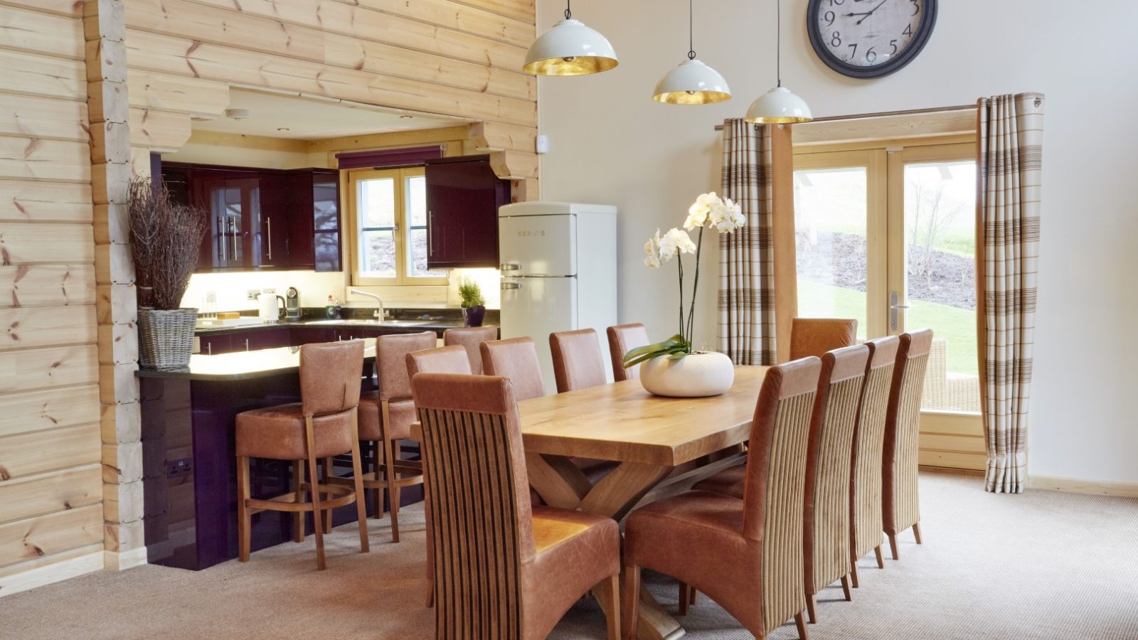 A spacious dining room at Celtic Manor features a large wooden table surrounded by brown upholstered chairs. Three pendant lights hang above, perfectly complementing the adjacent kitchen area with bar stools and a modern refrigerator. A clock and curtains decorate this bright Hunter Lodges space.