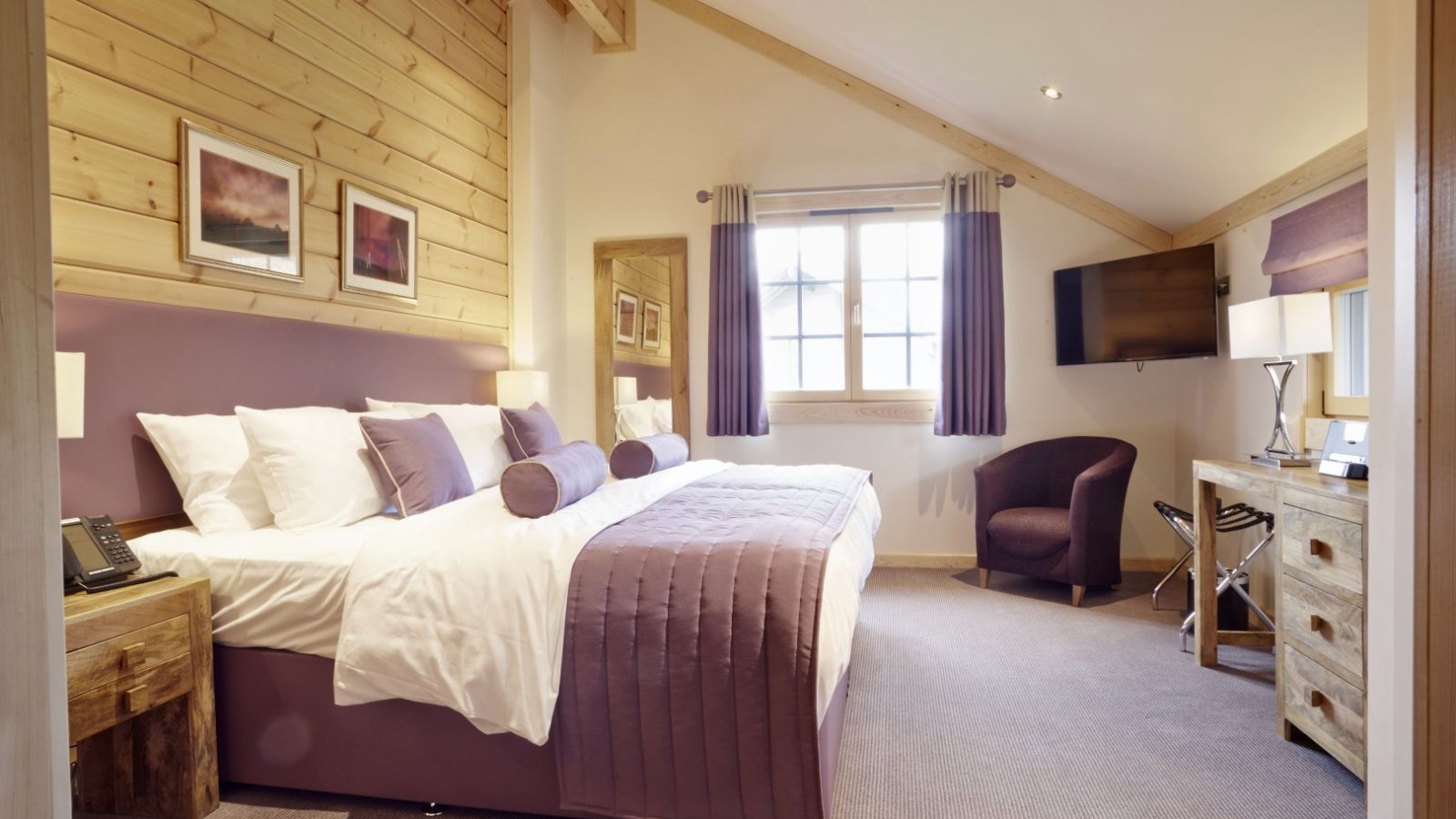 A cozy bedroom at the Hunter Lodges with a large bed featuring purple bedding and multiple pillows. The room boasts wooden walls, a sloped ceiling, and framed art. A TV is mounted on the wall alongside a small desk with a chair and an armchair by the window.