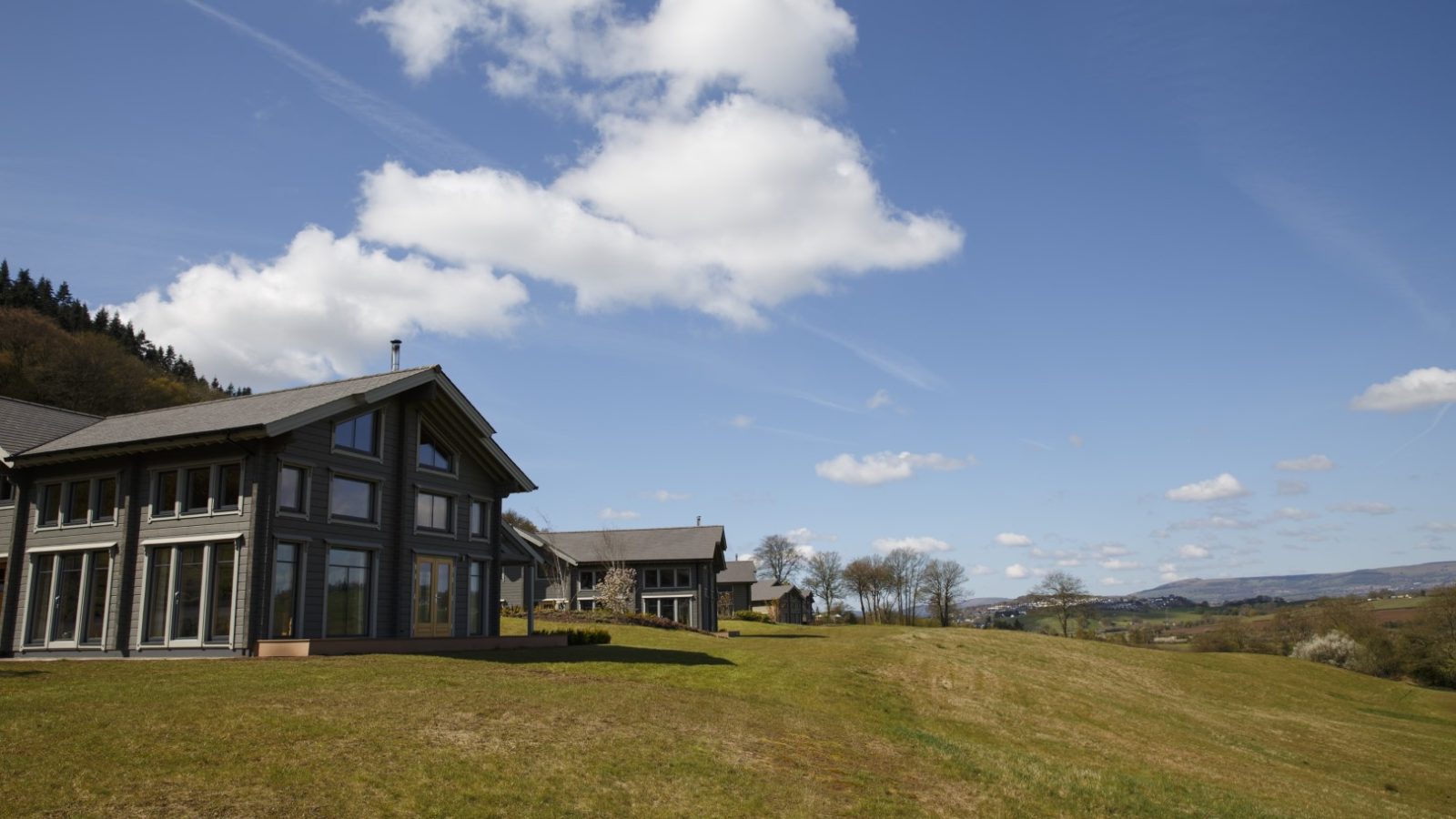 Perched on a grassy hill, a modern house with large windows stands under a clear blue sky with scattered clouds. Nearby, other homes dot the rolling hills and trees, evoking the serene charm of Hunter Lodges at Celtic Manor.