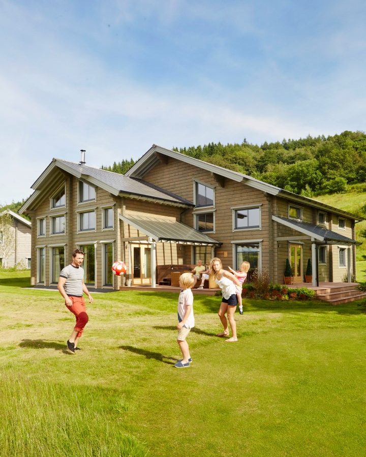 A family is playing on the grass in front of a large, modern wooden house reminiscent of the Hunter Lodges at Celtic Manor. The sky is clear, and the lush greenery envelops the area. Children are actively engaged in a game while adults bask in the sunny day.