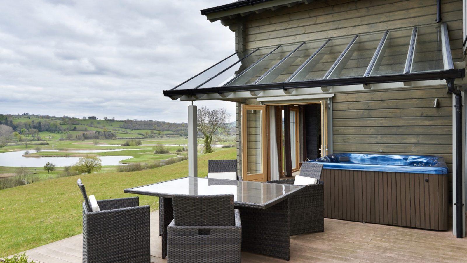 At Hunter Lodges, a wooden house with a glass veranda overlooks a lush green landscape and lake. On the patio, there is a hot tub and a wicker dining set with four chairs around a glass-top table. The sky is overcast, adding to the serene atmosphere of this Celtic Manor retreat.