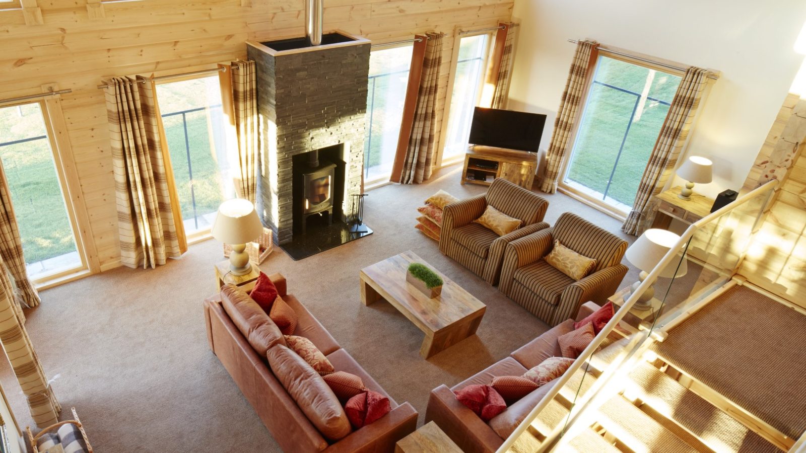 The spacious, sunlit living room of this Celtic Manor lodge features wooden walls with two sofas and armchairs around a coffee table. A modern fireplace sits against a black stone wall, and large windows with plaid curtains flood the space with natural light.