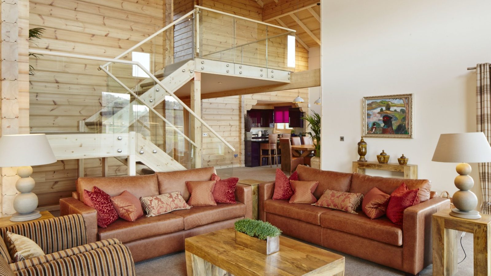 The living room at Hunter Lodges is a spacious haven with wooden walls and high ceilings, featuring two brown leather sofas with red cushions. A wooden coffee table sits center stage, while a glass-railed staircase ascends to a Celtic Manor loft adorned with art and decorative items on the walls.