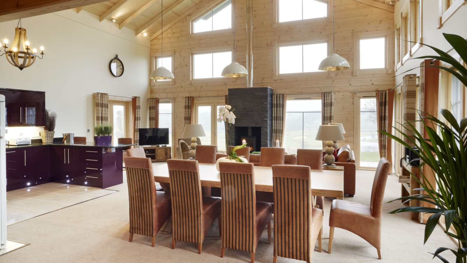 A spacious open-plan living and dining area in the style of Celtic Manor, featuring a wooden dining table with tall-backed chairs. The room boasts large windows, a high vaulted ceiling, and is adorned with plants and warm lighting. A kitchen from Hunter Lodges is visible in the background.