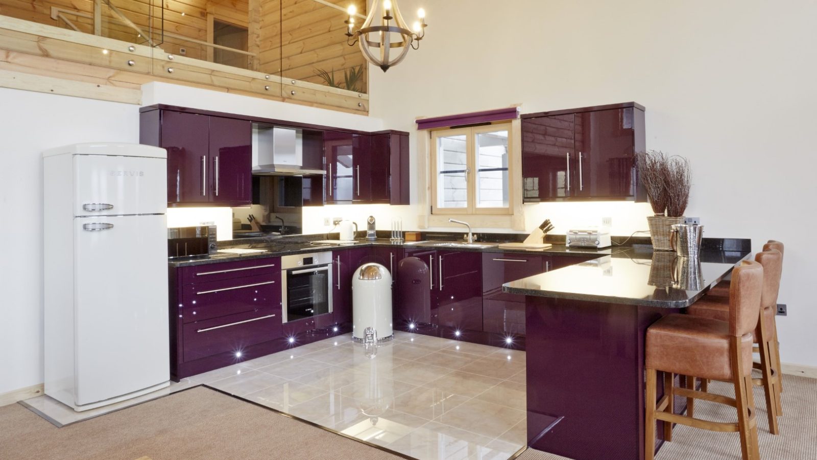 The modern kitchen, featuring glossy purple cabinets and black countertops, exudes the charm of a Celtic Manor. A vintage-style white refrigerator stands on the left, complemented by a wooden ceiling and upper-level balcony. Bar stools at the counter invite relaxation under soft, cozy lighting.