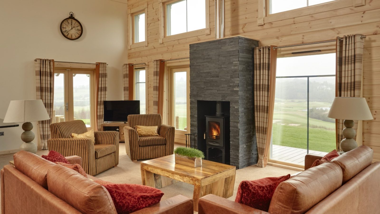 A cozy living room in a Hunter Lodges wooden cabin boasts large windows, a modern black fireplace, brown leather sofas, armchairs with red cushions, a wooden coffee table, and a wall clock—all with stunning views of the lush grassy landscape.