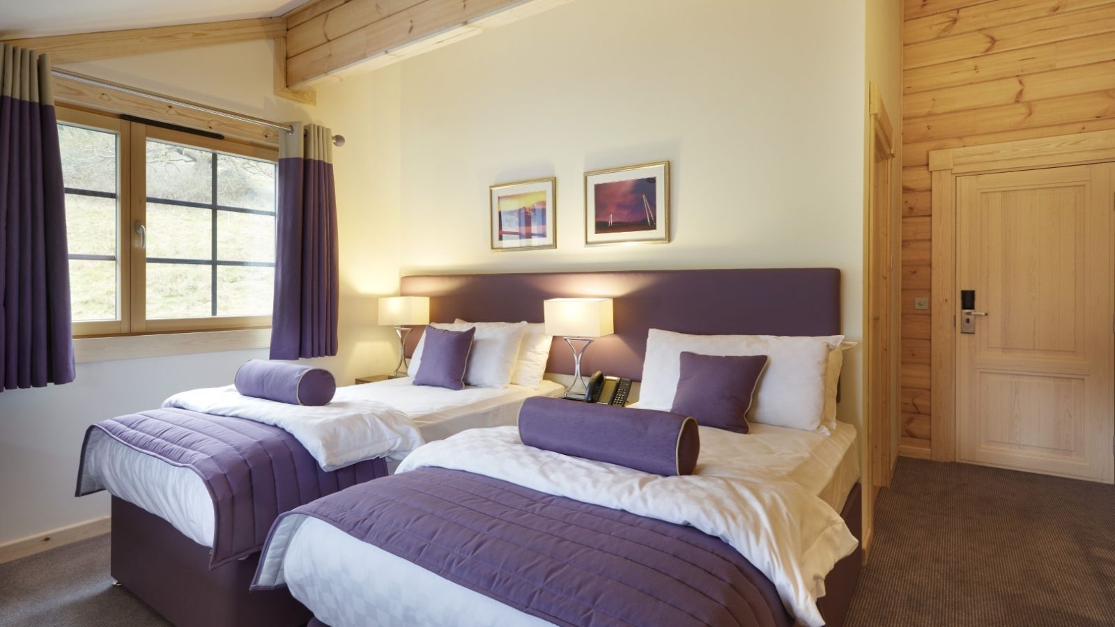 A cozy hotel room at Hunter Lodges features two double beds with purple bedding and pillows. The space boasts wooden accents, soft bedside lamp lighting, and two framed wall pictures. Natural light streams through the window, while a closed door sits to the right.