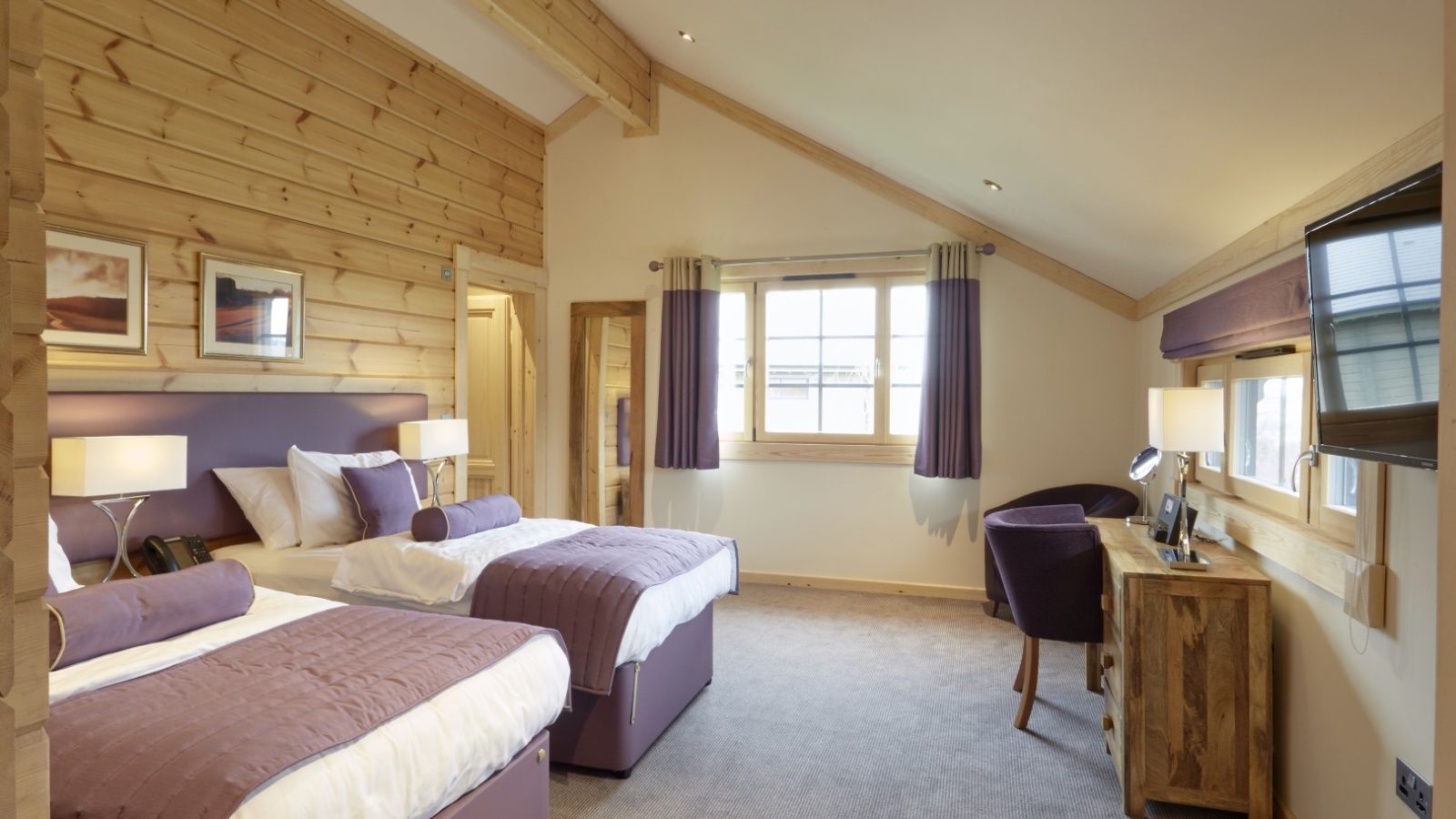 A cozy hotel room resembling the charm of Celtic Manor, with two double beds adorned in purple bedding and decorative pillows. The space features wooden walls, a small desk with a chair, a flat-screen TV, and a window with curtains welcoming natural light.