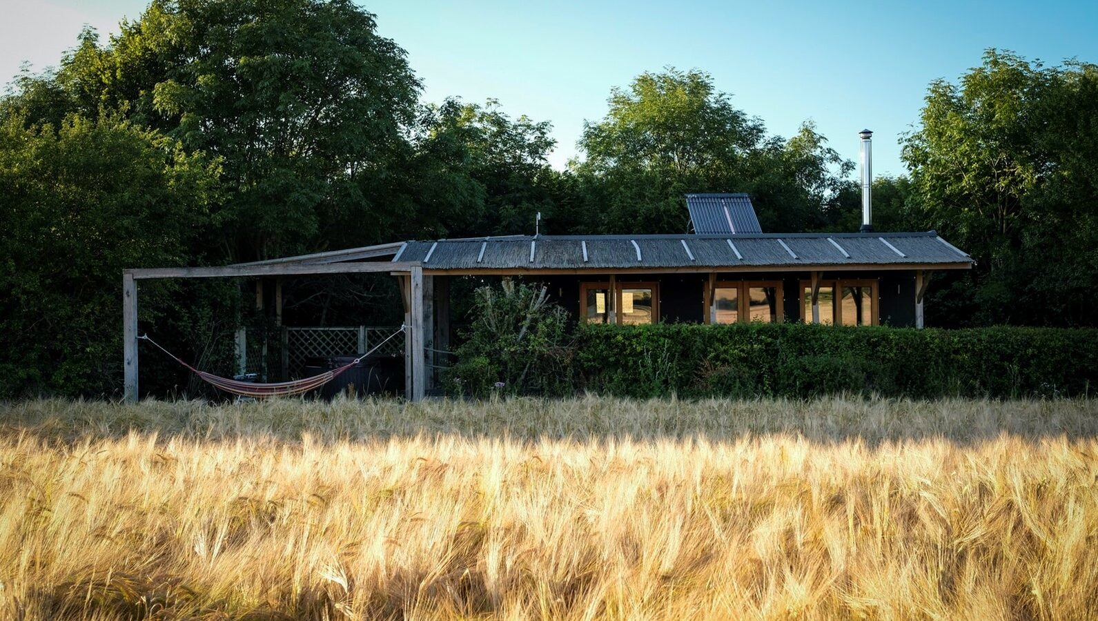 A small, single-story house nestled in the Chilterns features a hammock on the porch, surrounded by trees and a field of tall grass under a clear sky, offering a tranquil view.