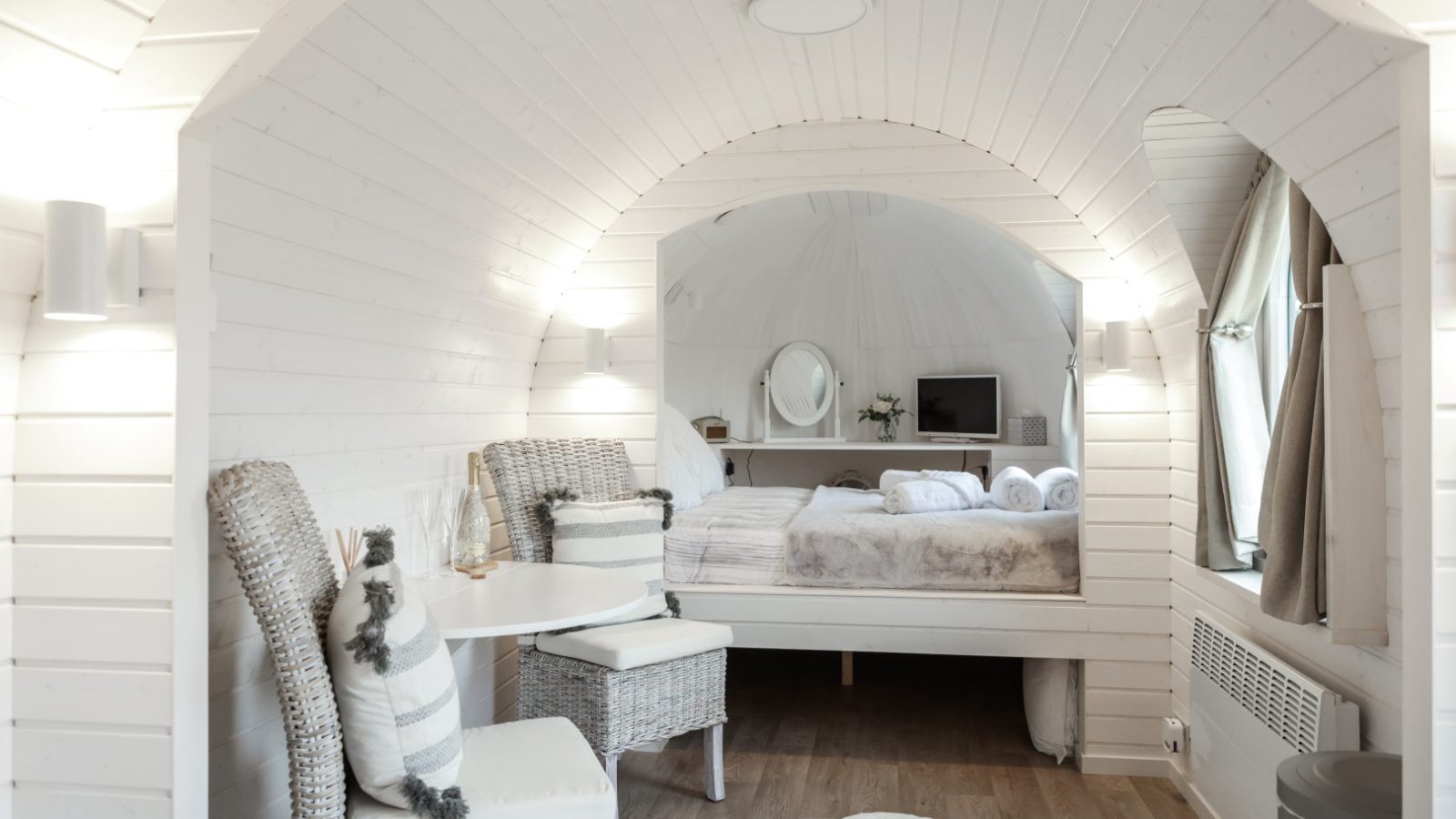 A cozy, white-painted wooden interior reminiscent of a castle chamber features a bed with fluffy bedding, a round mirror, and a small table with two wicker chairs. Soft lighting creates a warm atmosphere, while natural light escapes through the small window onto the fur rug on the wooden floor.